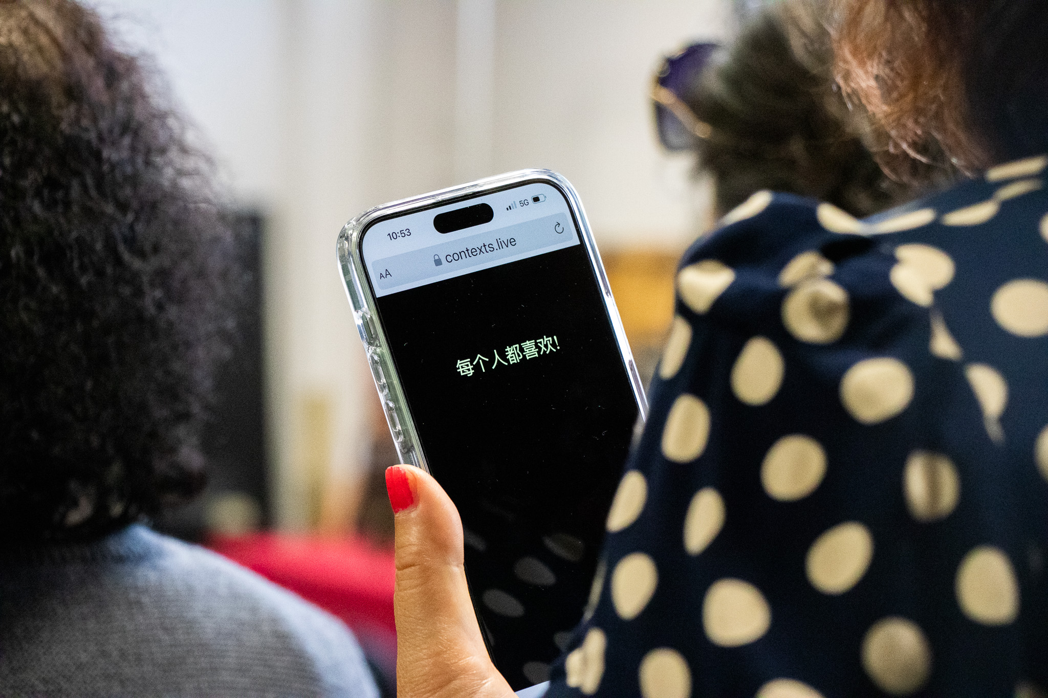 A woman holding an iPhone. On the screen is a web address of 'contexts.live' above a black background with one line of chinese characters on it.