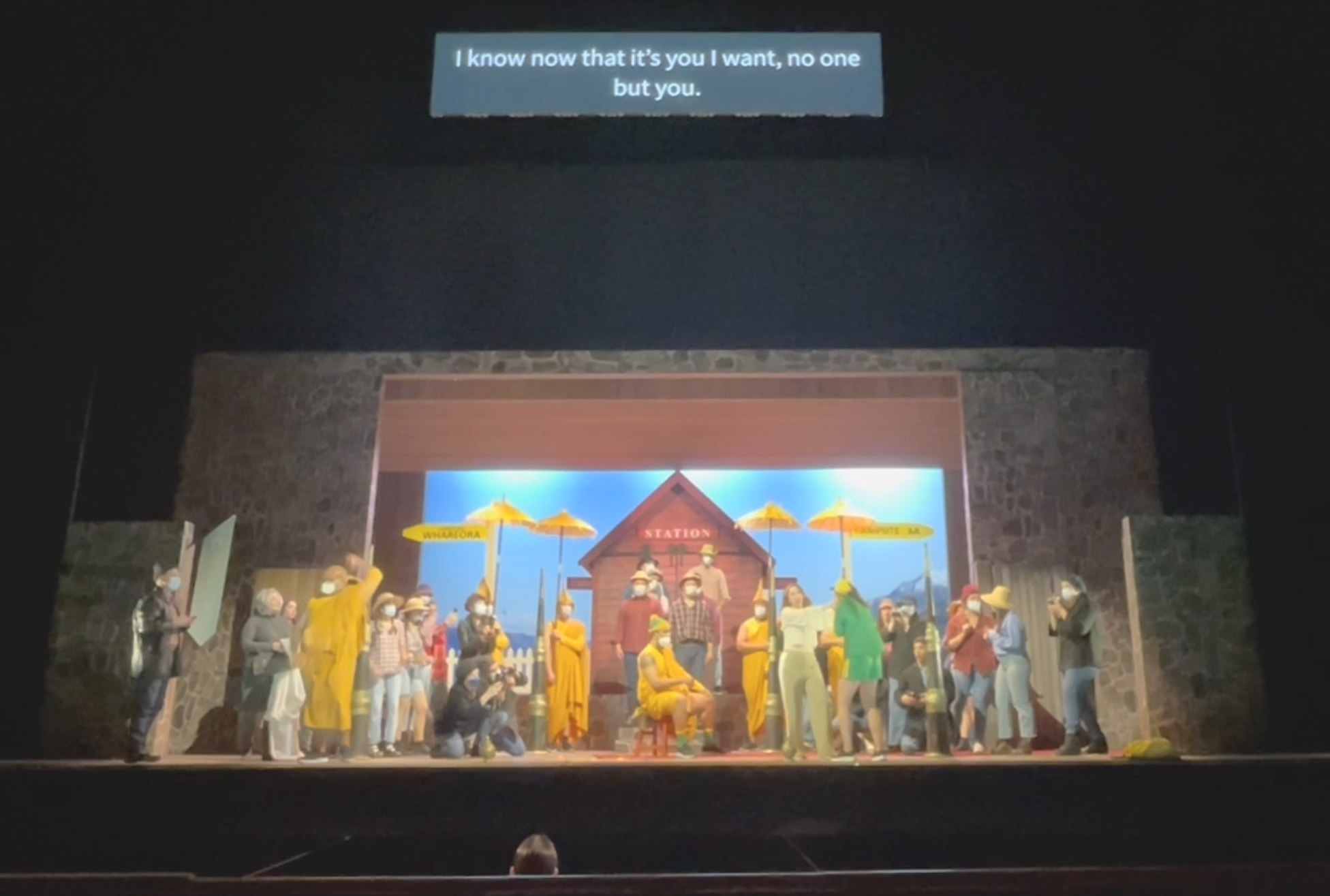 A brightly-lit stage in a darkened theatre, with many colourfully-dressed actors. Above the stage is a screen with words in English on it. In the foreground there is the back of a conductor's head, visible just above a screen.