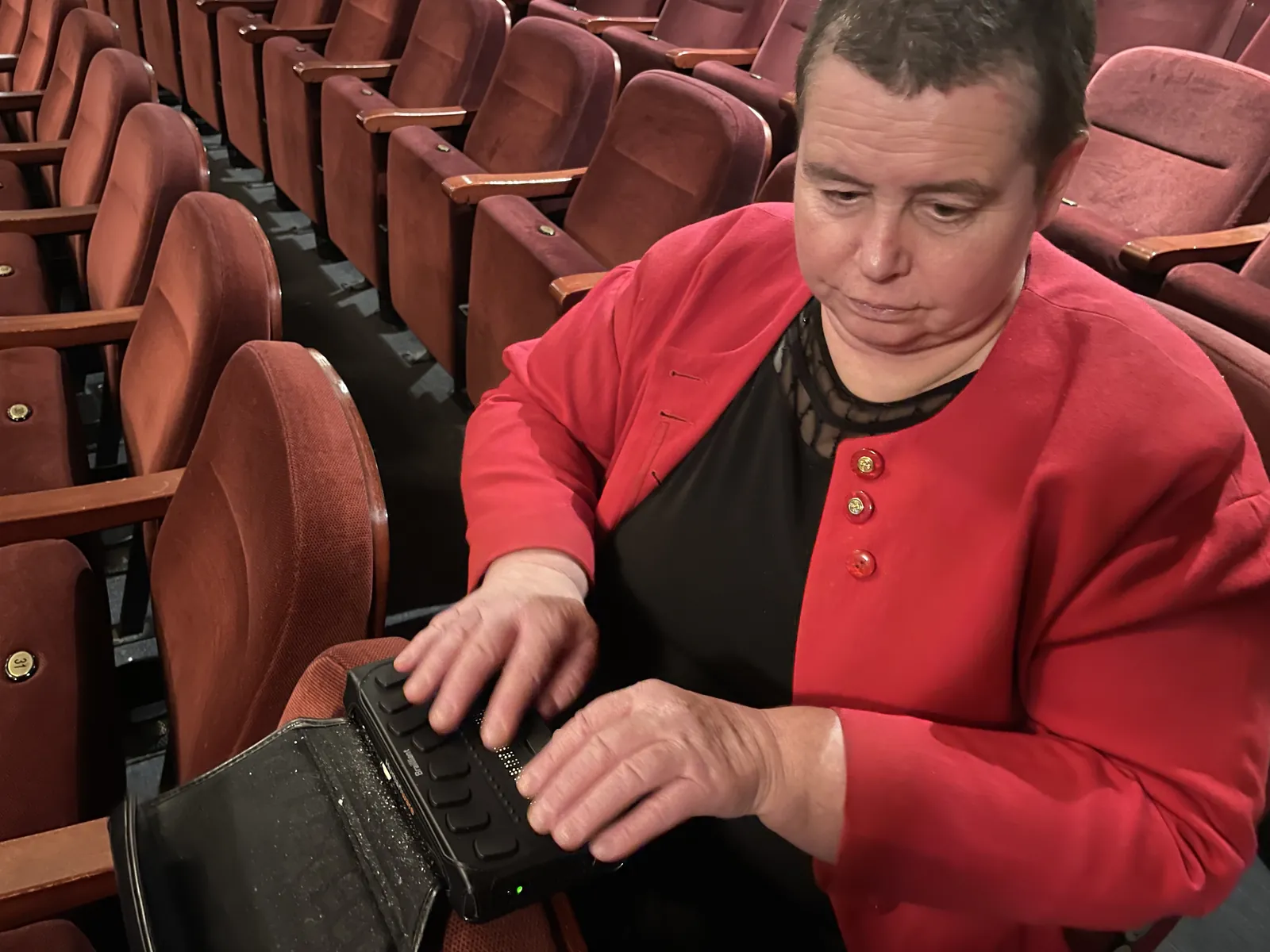 A woman in a red jacket using a portable braille display on the armrest at the opera