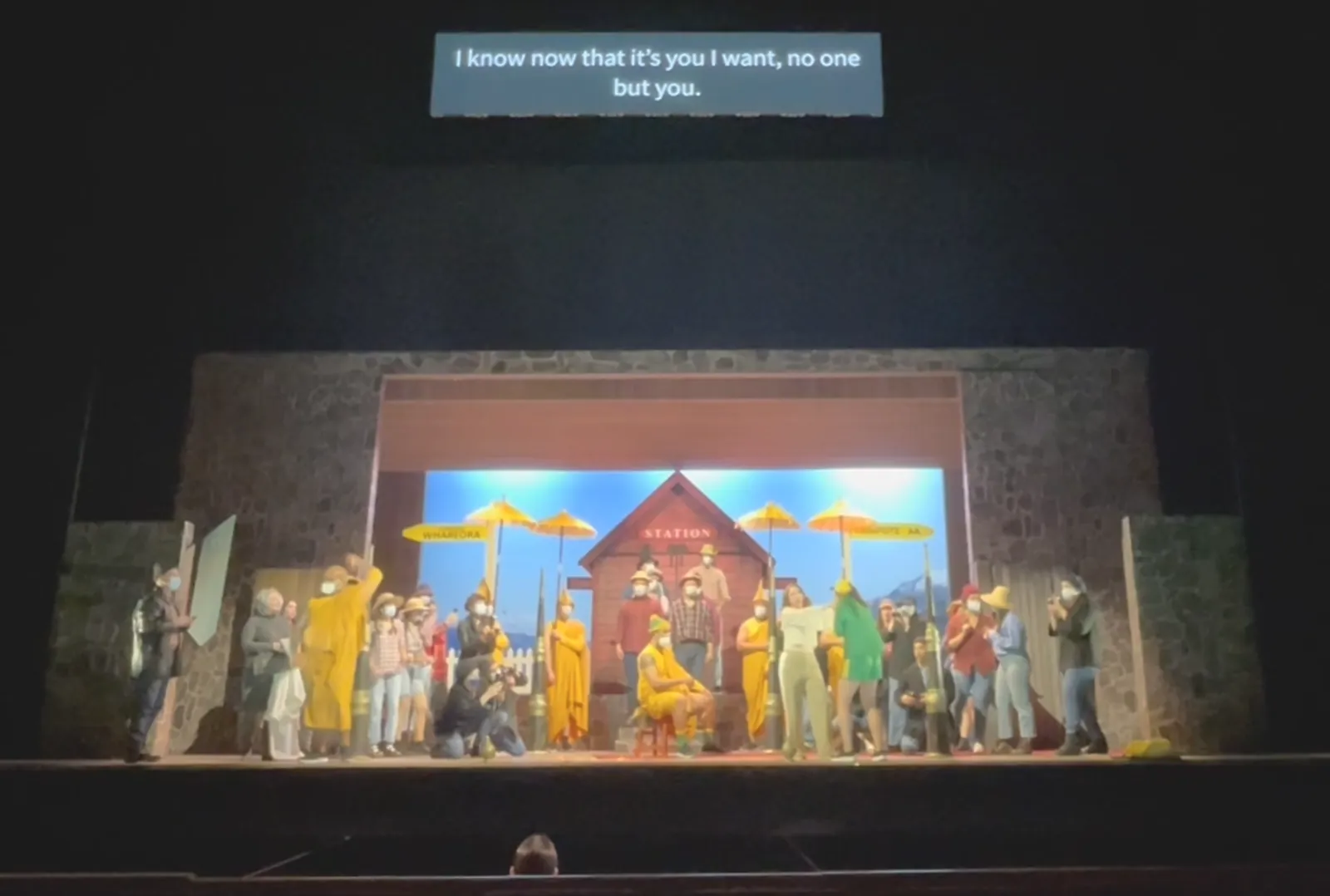 Stage performance with surtitles reading 'I know now that it's you I want, no one but you' — large ensemble cast in front of a painted backdrop