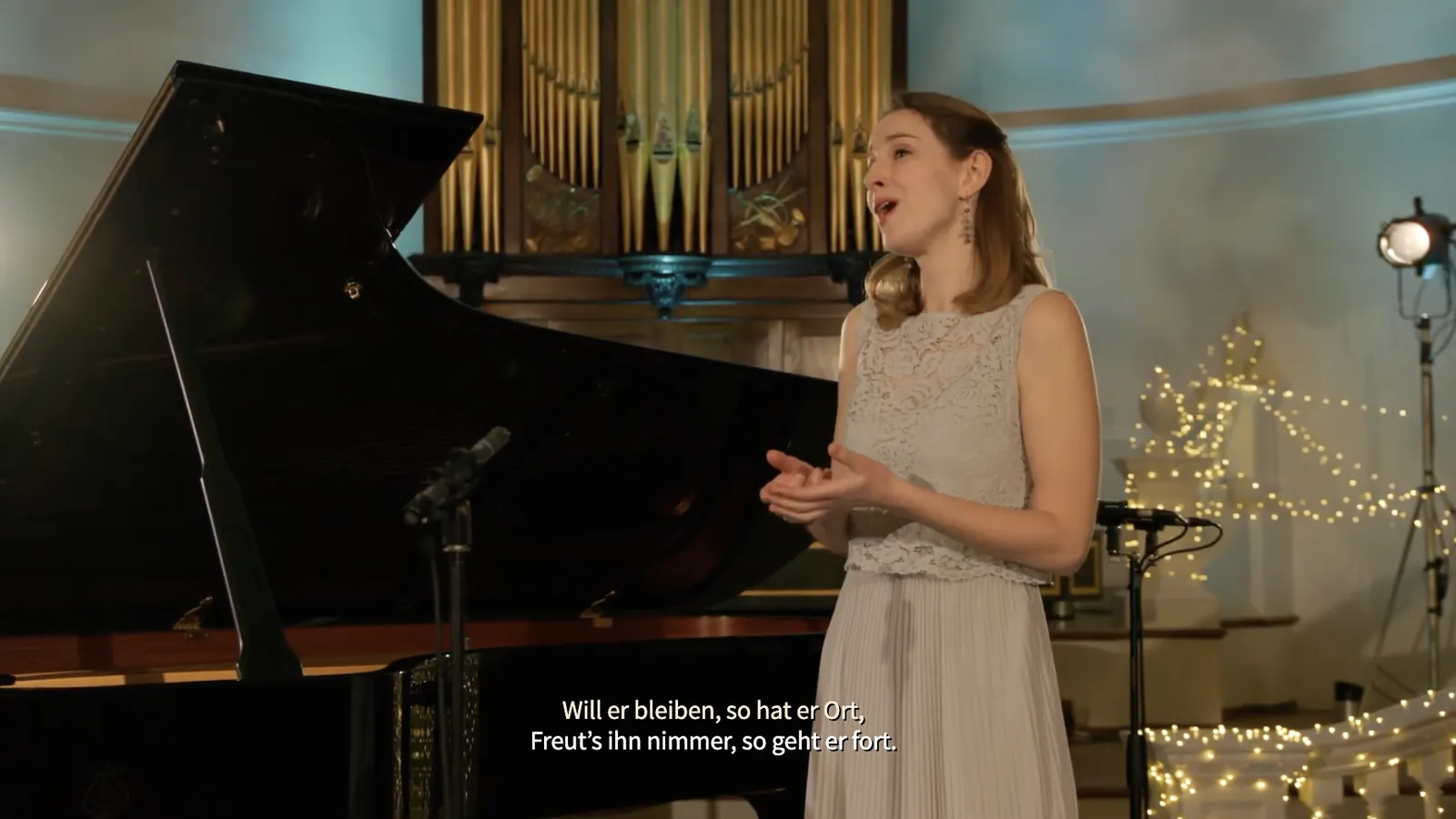 Singer beside grand piano, German surtitles at bottom